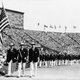 FILE - This is a July 29, 1948 file photo of Ralph Craig, center, of Albany, N.Y., who won the 100- and 200-meter dashes in the 1912 Olympic games in Stockholm, and is a member of the U.S. Olympic Yachting team in the 1948 games, carrying the American flag in the parade of the nations at the opening of the Olympic games in London's Wembley Stadium. Athletes bought their own uniforms, and some their own food. They stayed in private homes, schools and military barracks. If eggs appeared on the training menu, it was a cause for celebration. When London hosted the Olympics in 1948, organizers did it on the cheap, and they made no apologies about it. (AP Photo/File)